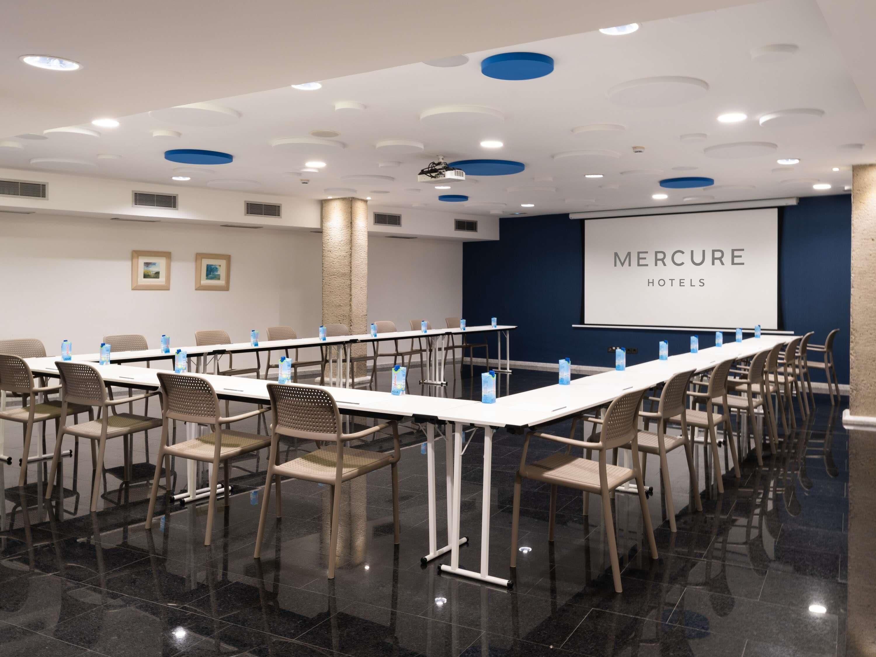 Un salón de conferencias con una larga mesa, sillas, botellas de agua y una pantalla de proyector, con un cartel de 'Mercure Hotels'.