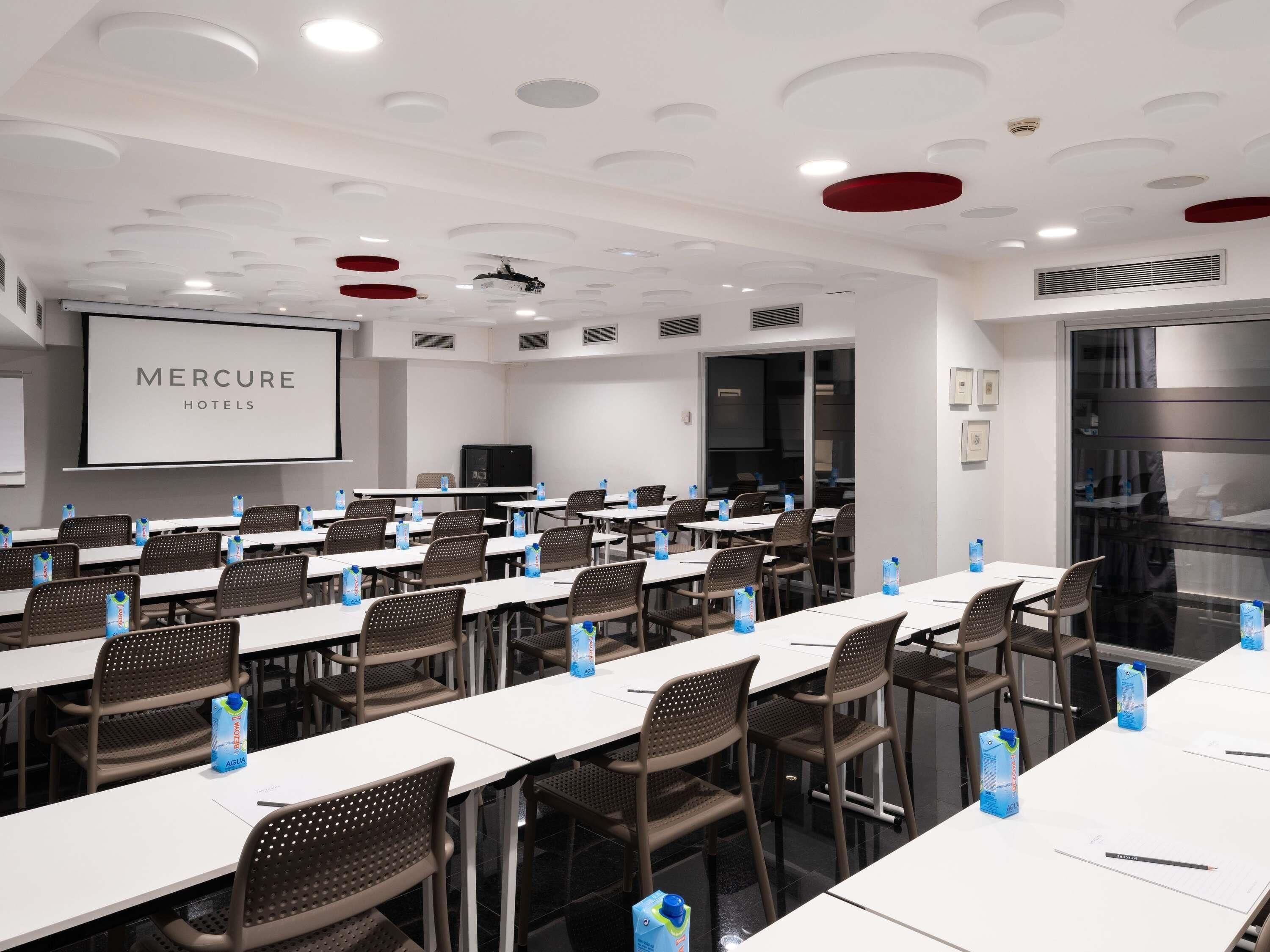 Una sala de conferències amb taules blanques, cadires marrons i una pantalla projectant 'MERCURE HOTELS'.