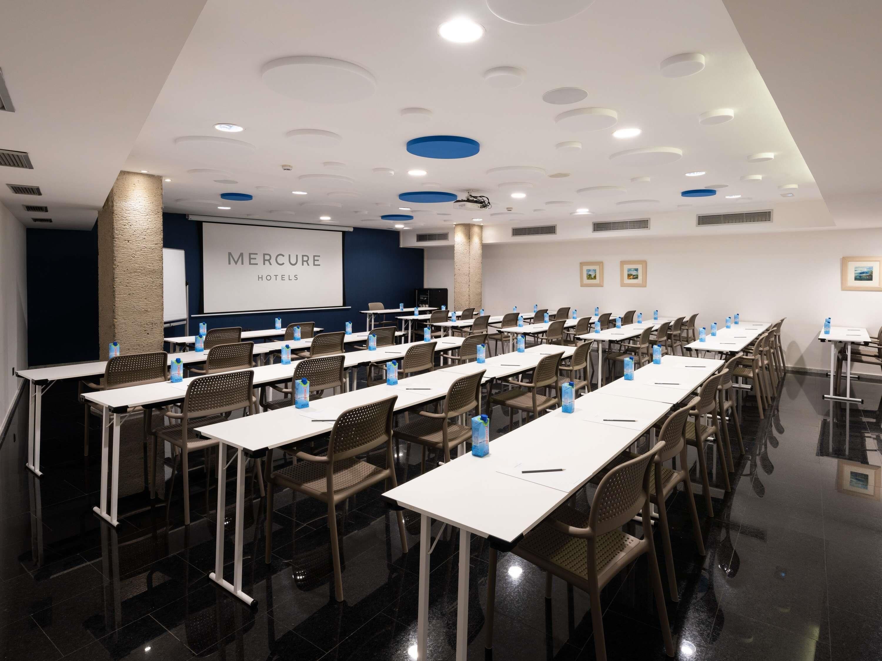 Un salón de conferencias con mesas, sillas y una pantalla de proyección que muestra 'MERCURE HOTELS'.