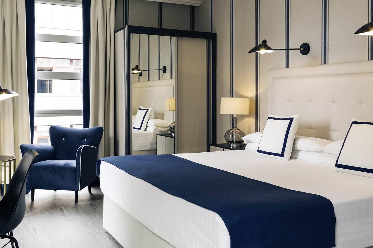 A hotel room with a large bed, a chair, a nightstand, a lamp, and a mirror.