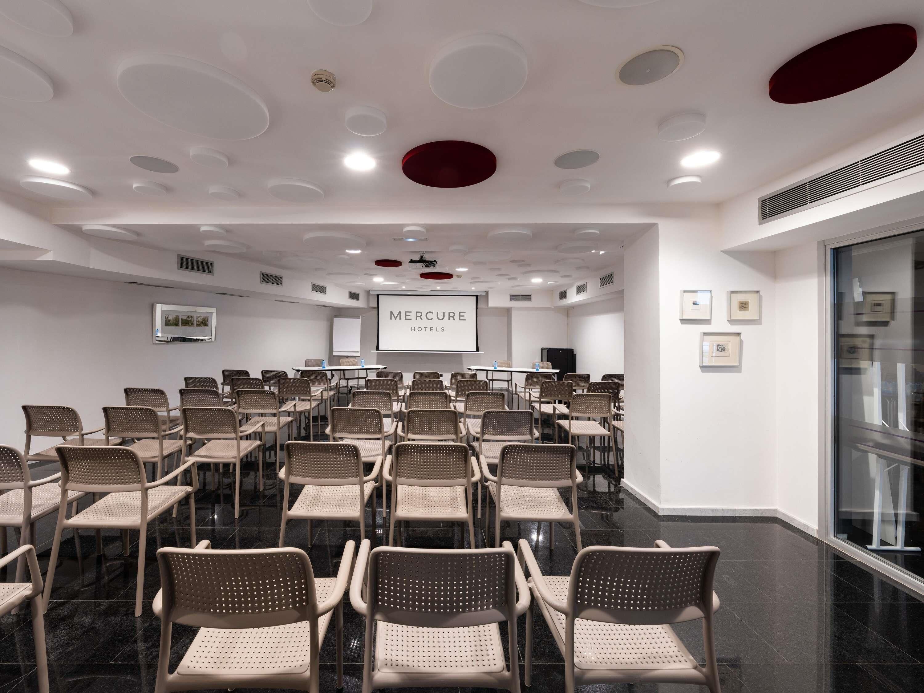 A conference room with a white ceiling, a large screen, and a sign reading 'Mercure Hotels'.