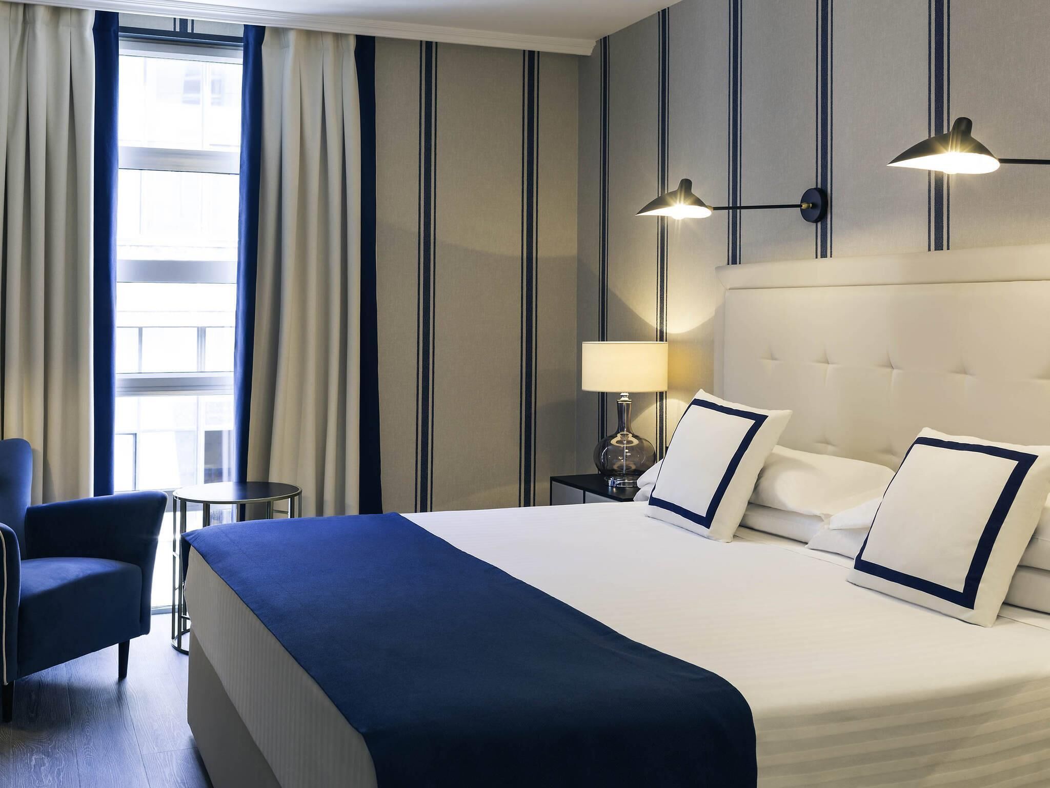 Habitación de hotel con cama, sillón, mesita de noche, lámpara y vistas al exterior por una ventana.