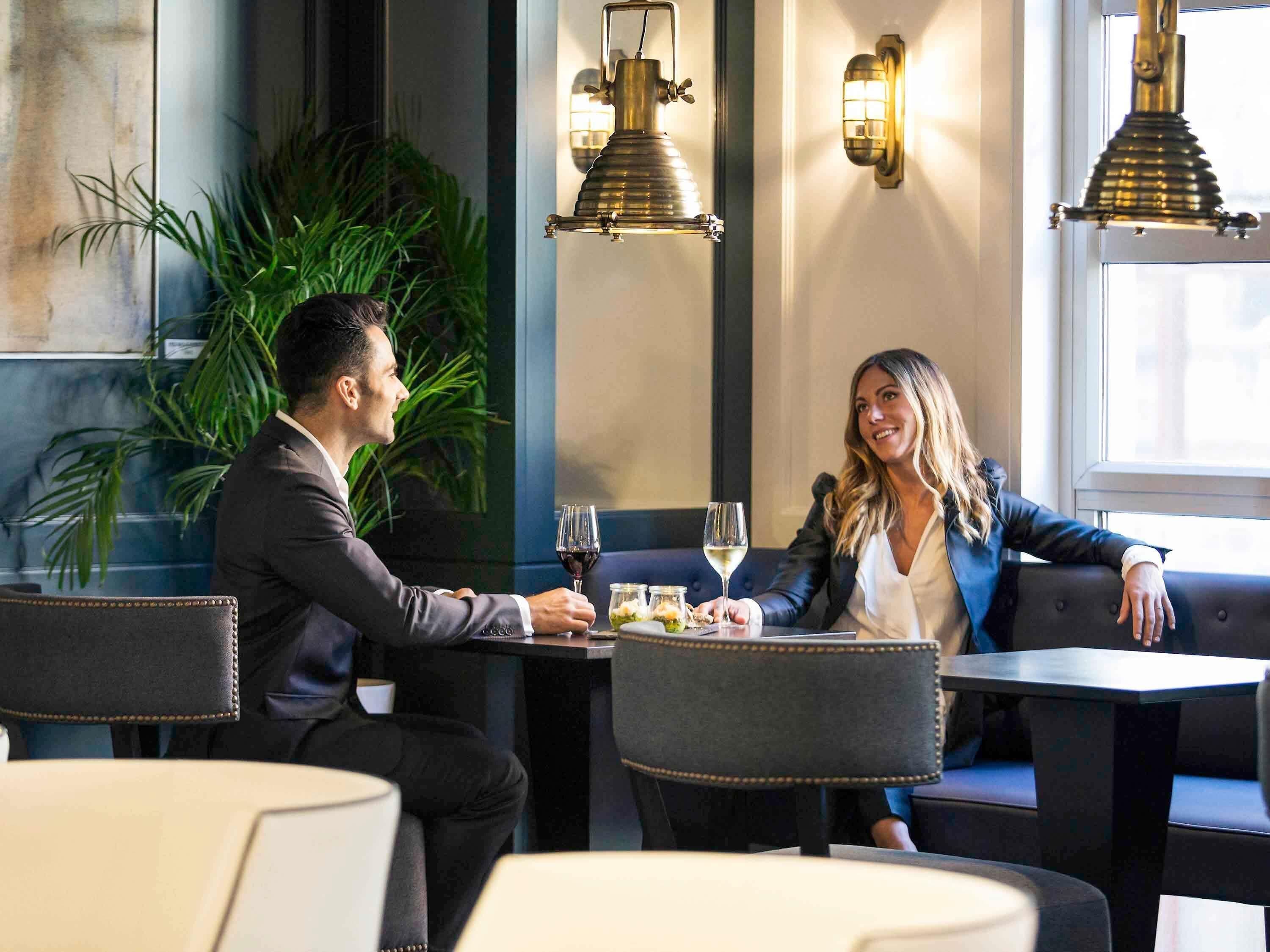 Un hombre y una mujer cenando en un restaurante con una decoración moderna, que incluye una planta, lámparas colgantes y vistas de la ciudad.