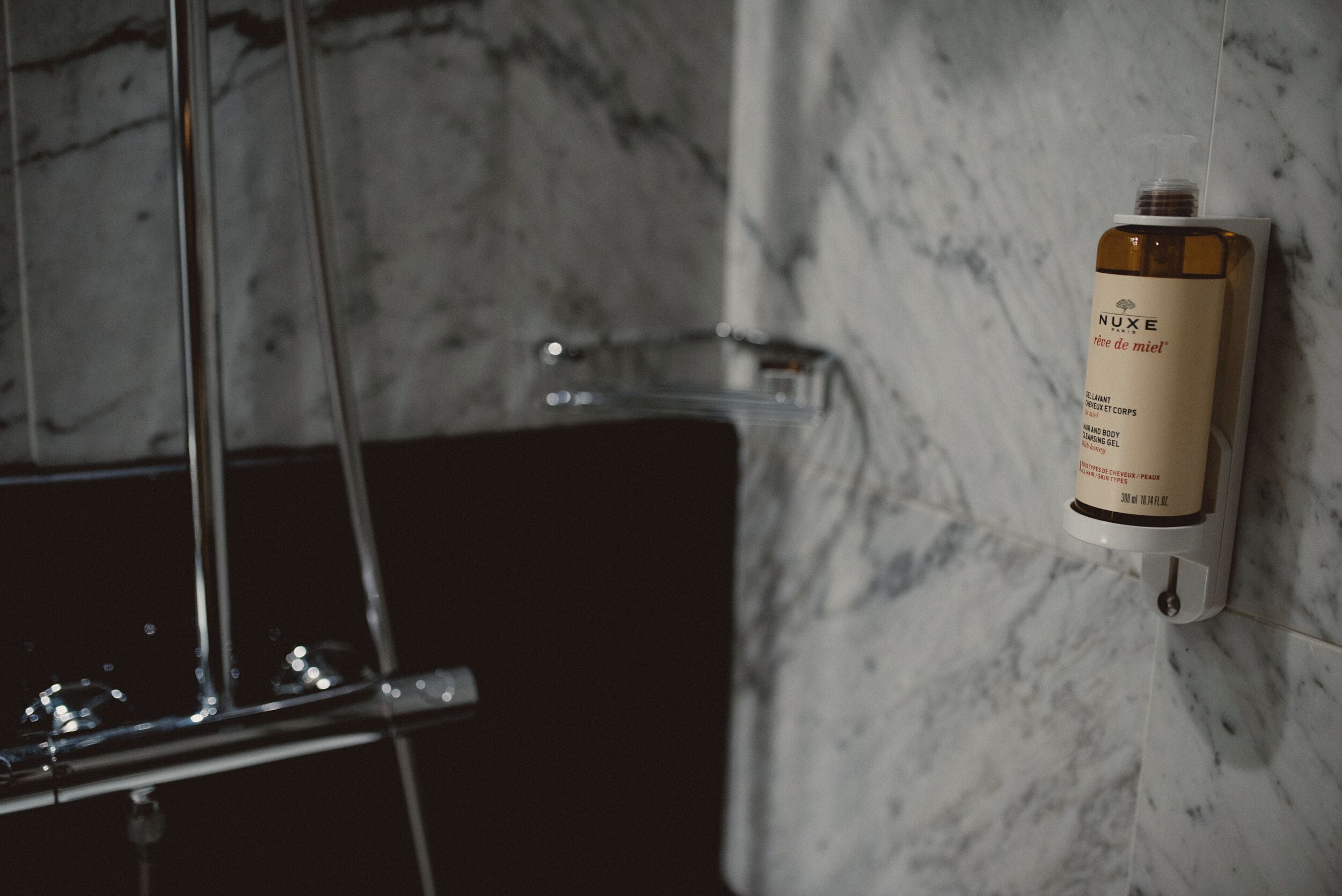 A bathroom with a marble wall, a shower enclosure, and a Nuxe body lotion dispenser.