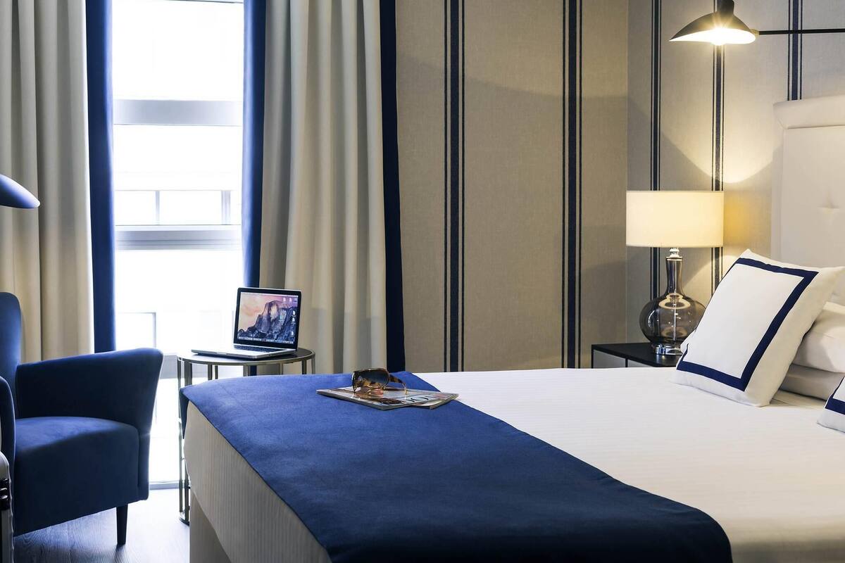 A hotel room with a bed, a desk, a chair, a lamp, and a window with curtains.
