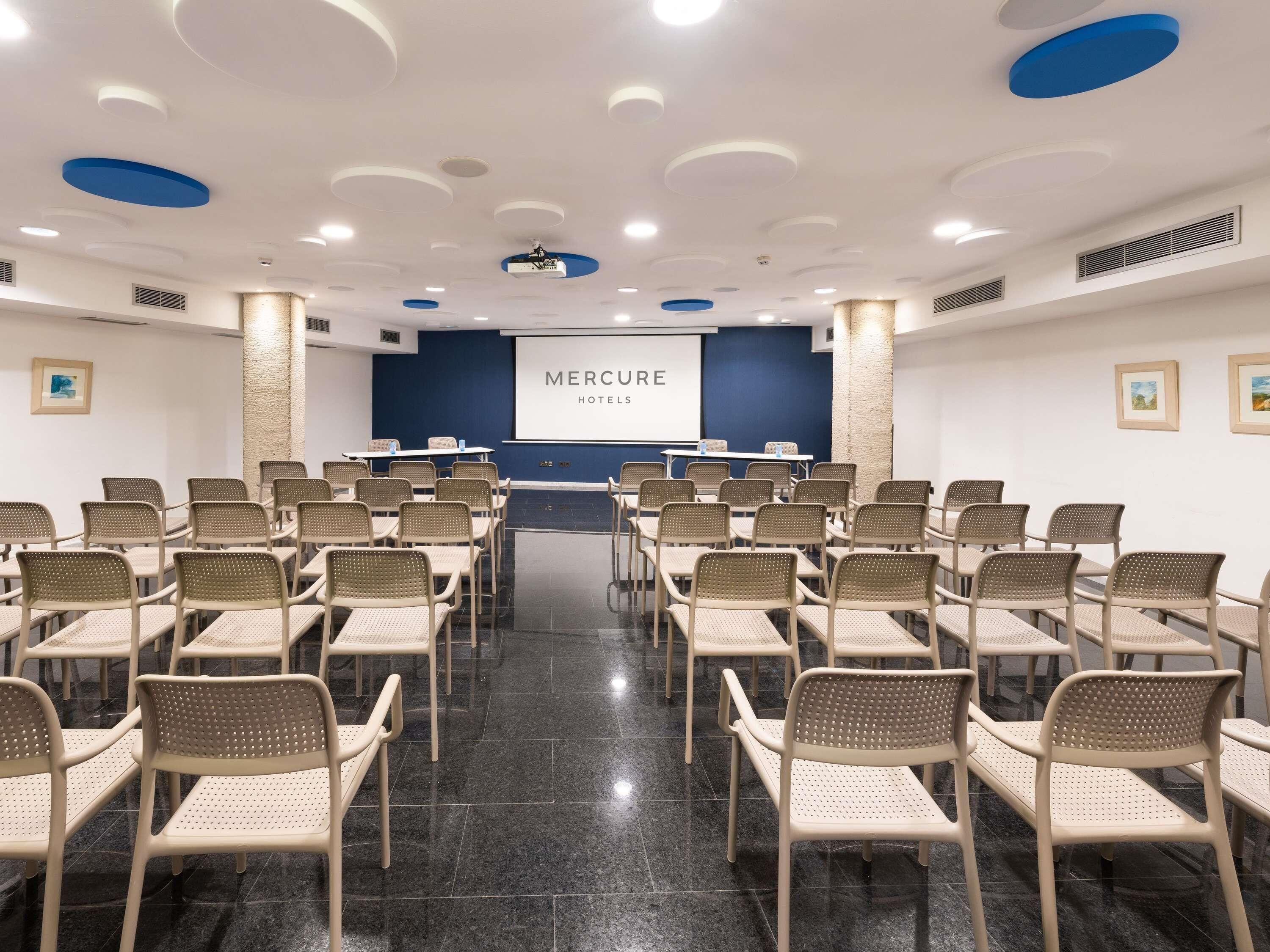 A conference room with rows of beige chairs, a projector screen displaying 'MERCURE HOTELS', and a blue accent wall.
