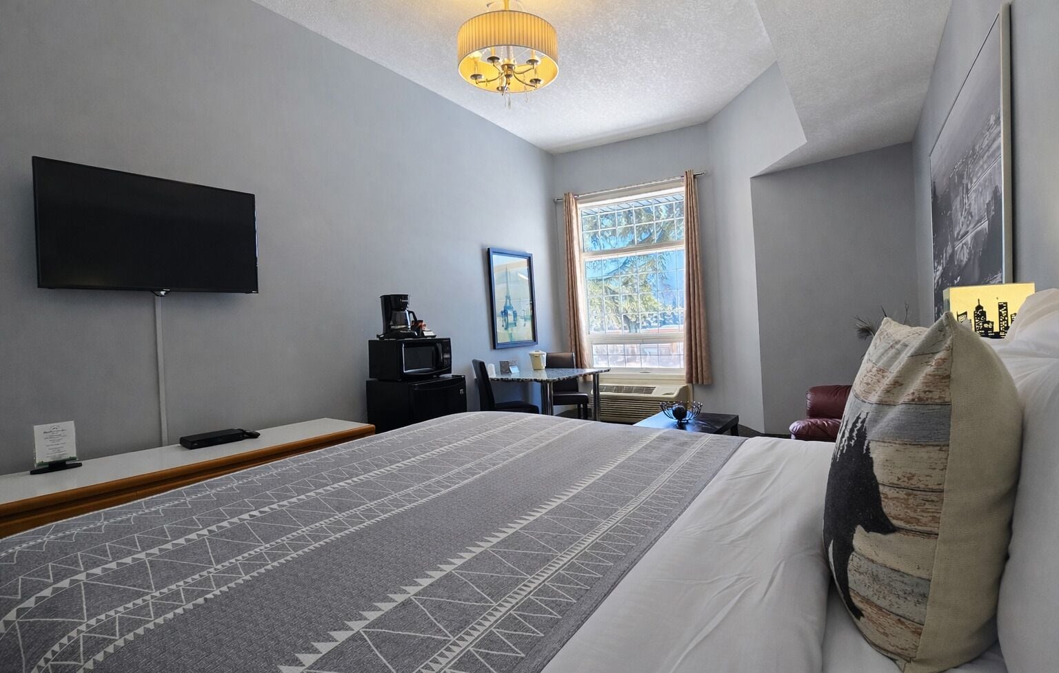 A hotel room with a large bed, a flat-screen TV mounted on the wall, a desk with a chair, and a window with curtains.