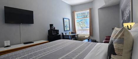A hotel room with a large bed, a flat-screen TV mounted on the wall, a desk with a chair, and a window with curtains.