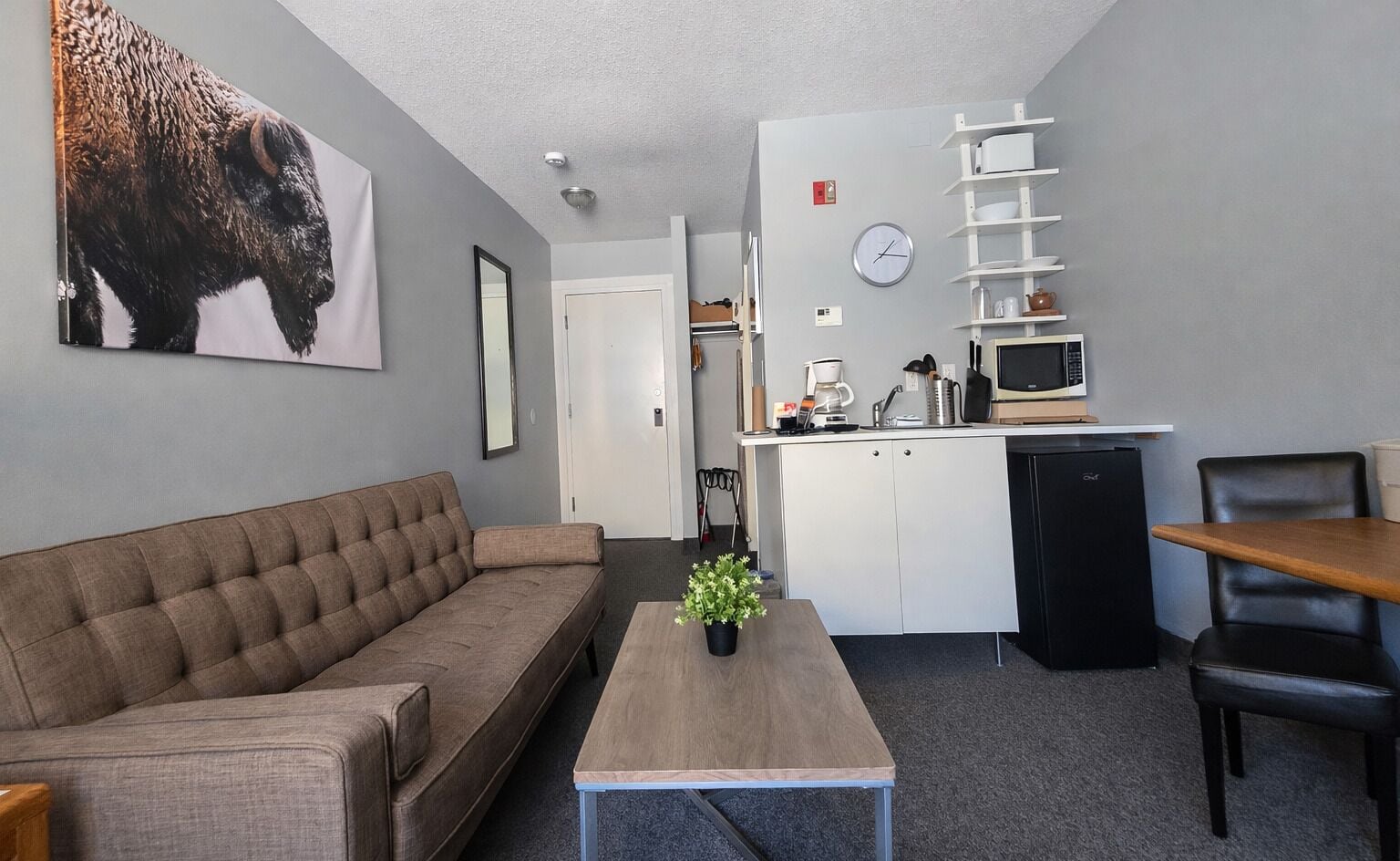 Een moderne woonkamer met een bank, een salontafel en een eethoek met een tafel en stoelen. Er is een kitchenette met een magnetron, koffiezetapparaat en kastjes. Een klok en een ingelijst kunstwerk hangen aan de muur.