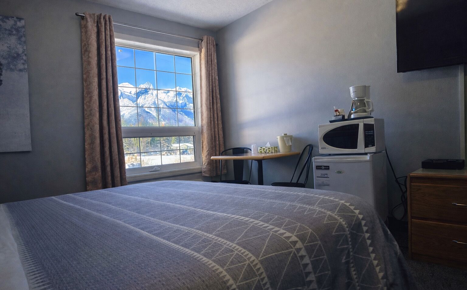 Habitación de hotel con cama, ventana con cortinas, escritorio con silla, microondas y televisor en la pared.