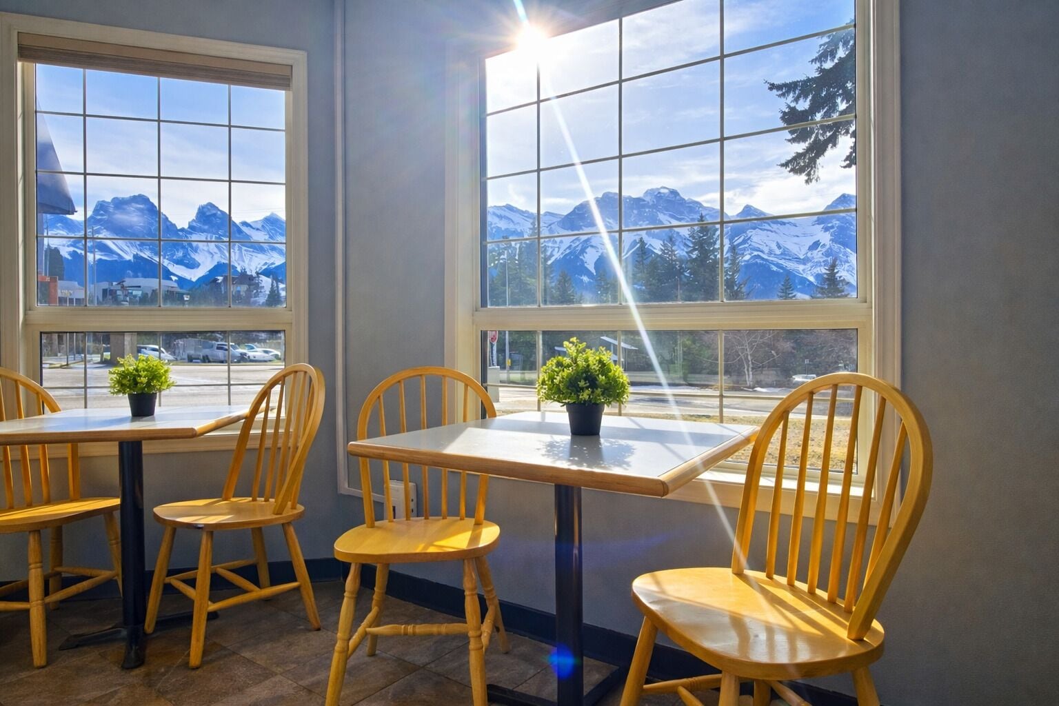 Une pièce avec deux tables et quatre chaises en bois, chacune agrémentée d’une petite plante en pot. De grandes fenêtres offrent une vue sur des montagnes enneigées.