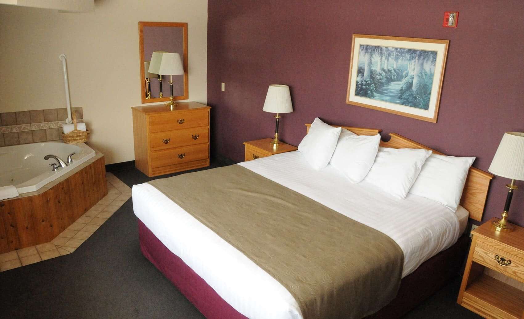 A hotel room with a large bed, a wooden dresser, a nightstand with a lamp, a bathroom with a bathtub, and a framed picture on the wall.