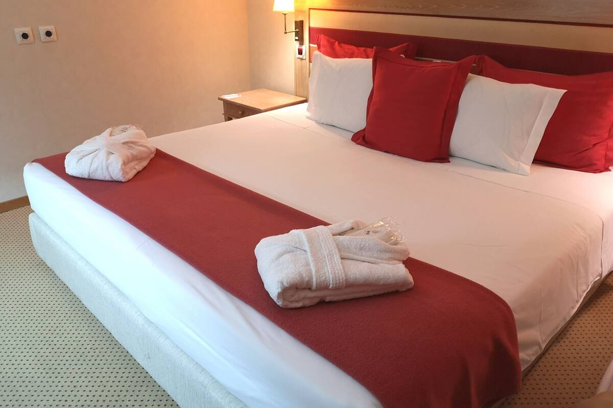 A neatly made bed with red and white bedding, two folded towels on the bed, a nightstand with a lamp, and a wall with two light switches.