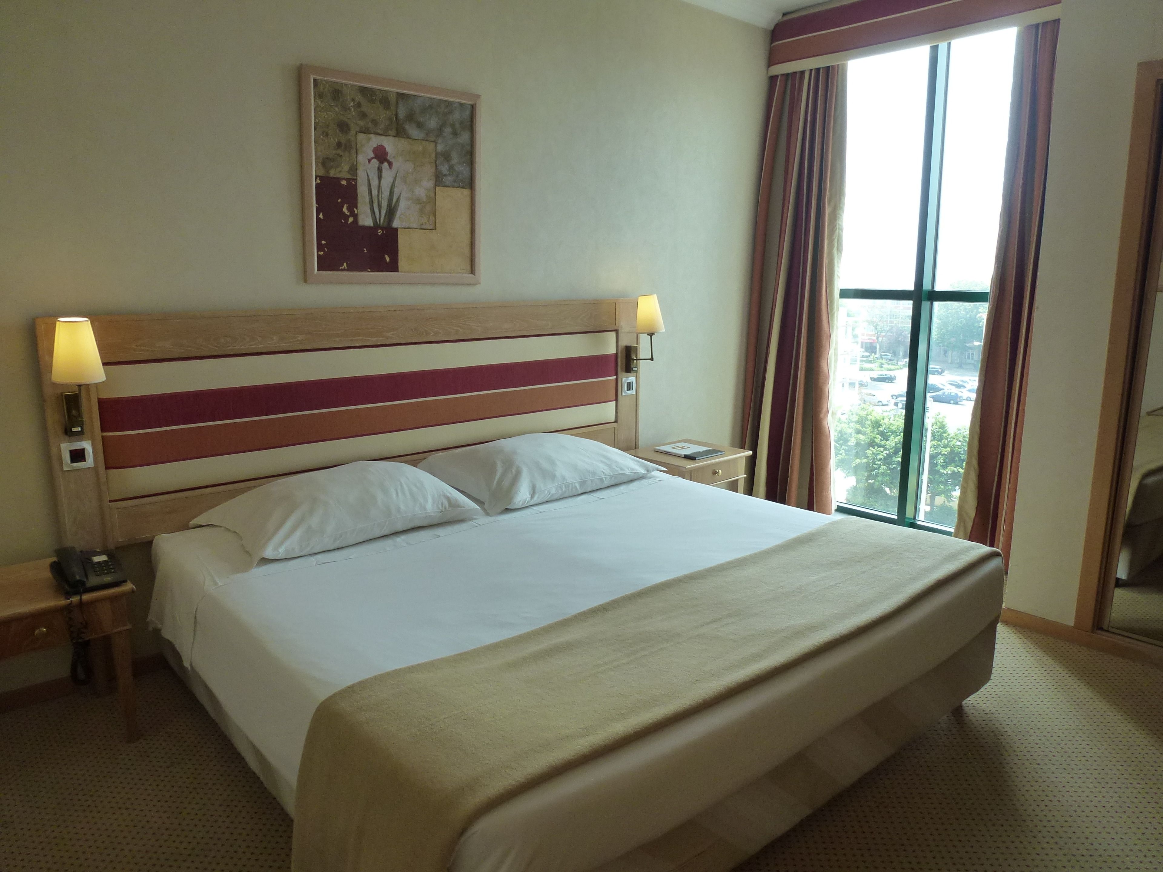 Ein Hotelzimmer mit einem großen Bett, zwei Nachttischlampen, einem Nachttisch, einem Telefon und einem Fenster mit Vorhängen.