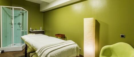 A room with a bed, a green wall, a glass shower enclosure, a standing lamp, and a green chair.
