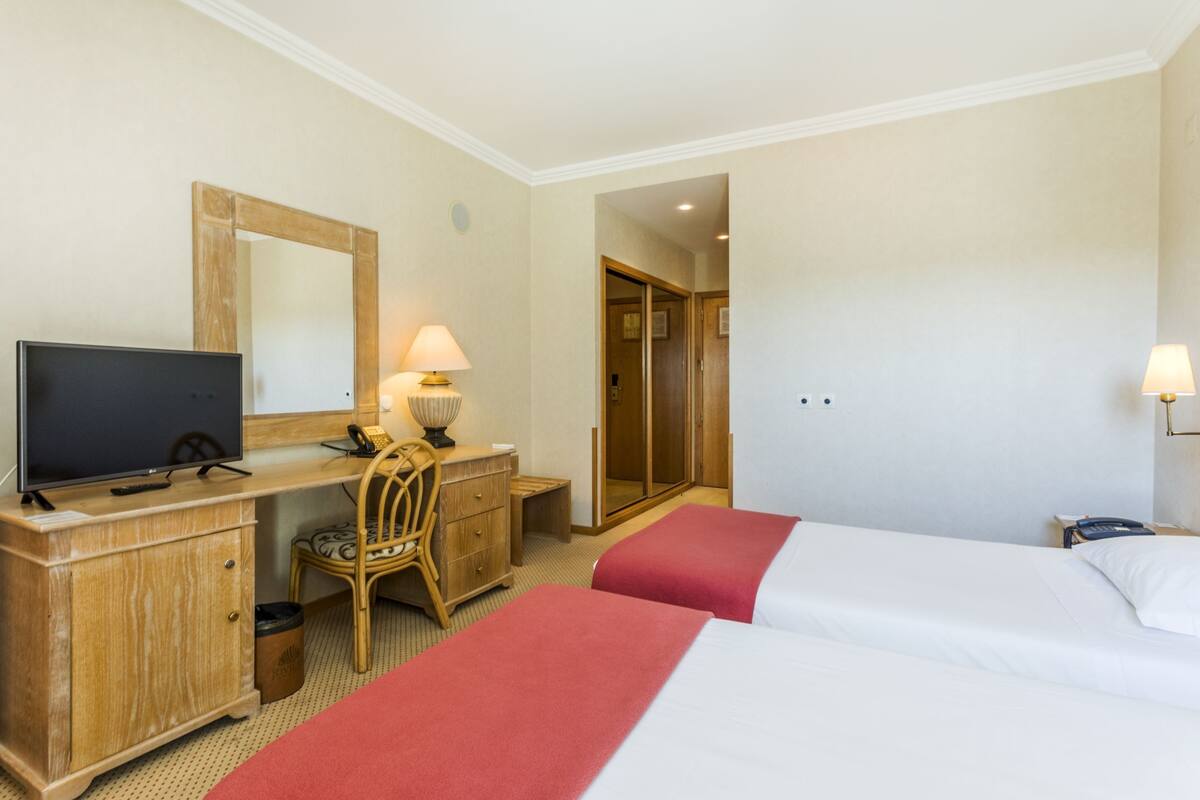 A hotel room with a TV, desk, chair, two beds, and a mirror.