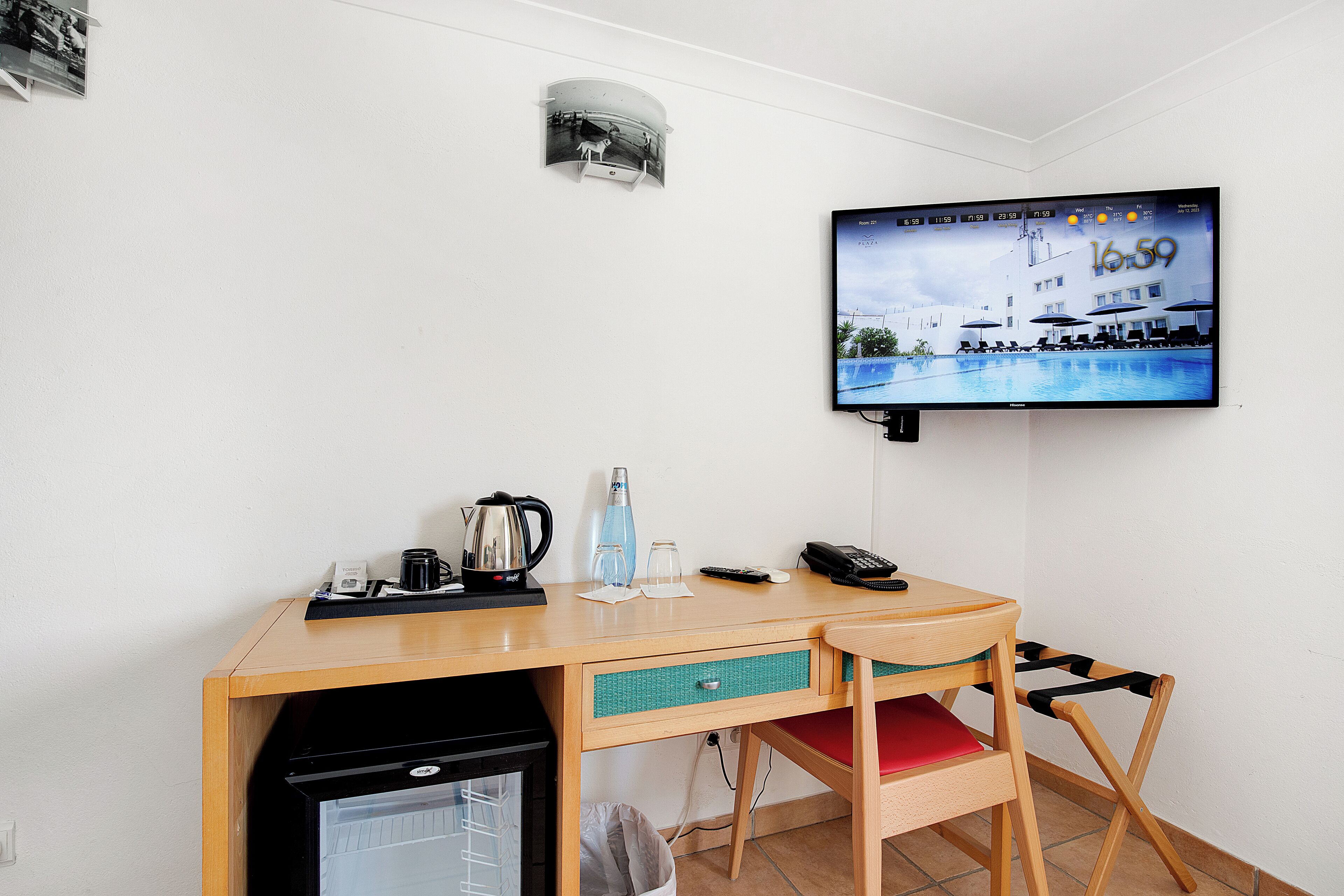 Habitación de hotel con un escritorio de madera, un televisor montado en la pared mostrando una escena de piscina, un refrigerador pequeño, una silla y un cesto de basura.