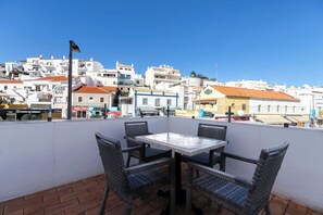 Une terrasse dotée de chaises et d’une table en osier, offrant une vue sur une ville aux bâtiments blancs et sous un ciel d’un bleu éclatant.