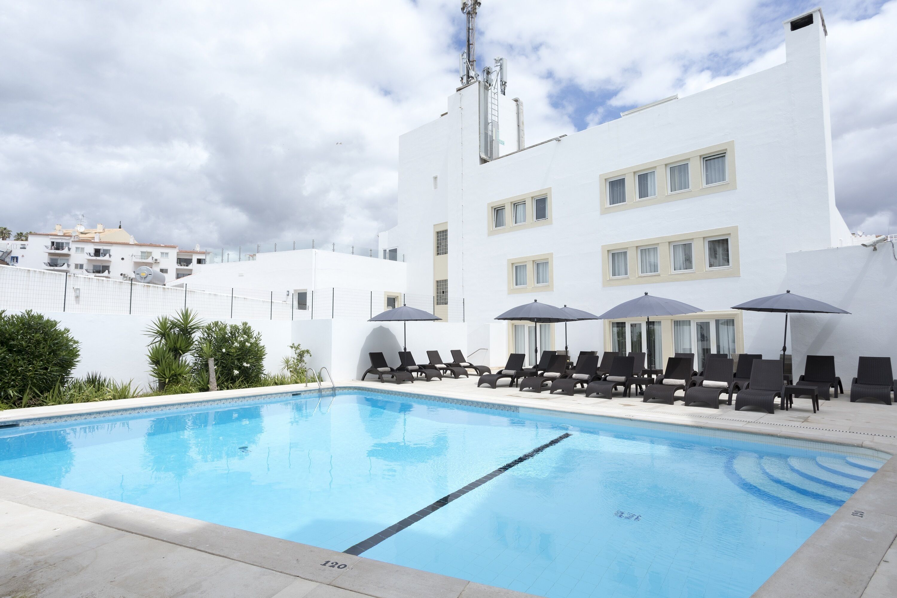 Ein Swimmingpool mit Liegestühlen und Sonnenschirmen, umgeben von einem weißen Gebäude mit zahlreichen Fenstern und einer flachen Dachkonstruktion.