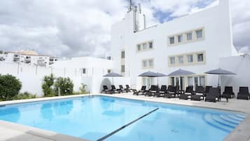 A swimming pool with lounge chairs and umbrellas, surrounded by a white building with multiple windows and a flat roof.