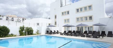 Ein Swimmingpool mit Liegestühlen und Sonnenschirmen, umgeben von einem weißen Gebäude mit zahlreichen Fenstern und einer flachen Dachkonstruktion.