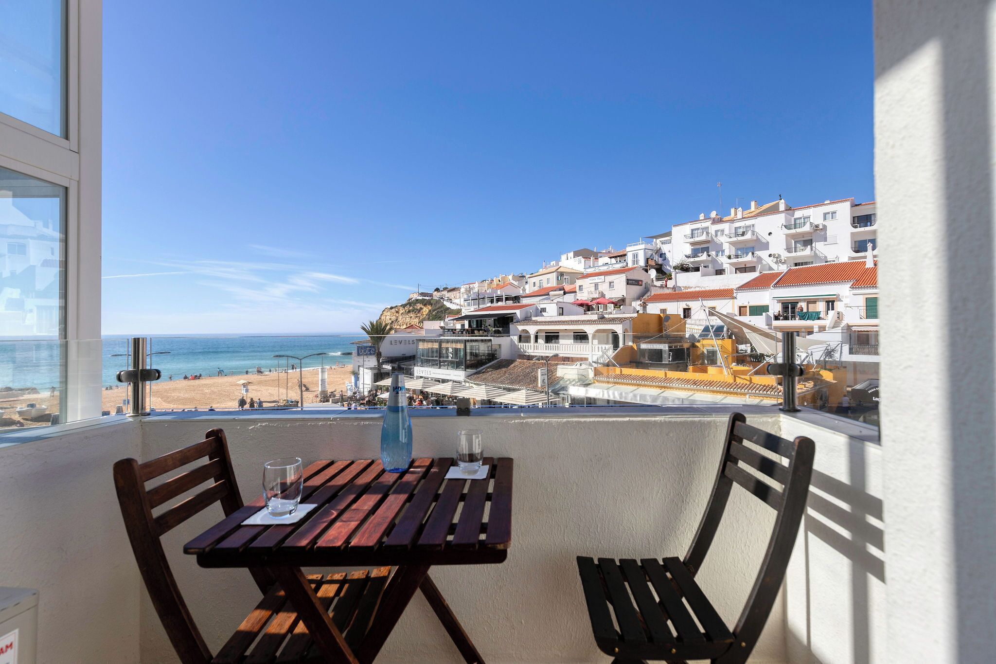Un balcon avec une table et des chaises donnant sur une plage et une ville située sur une colline.