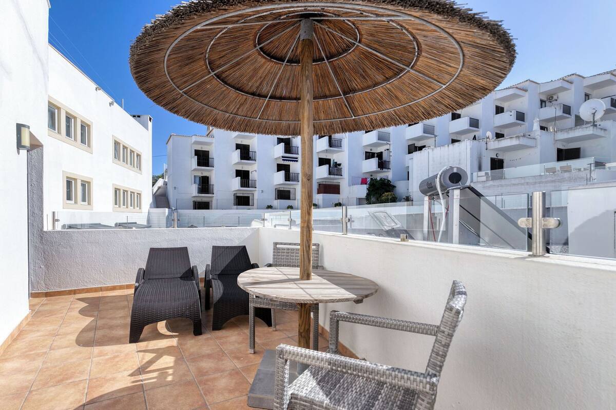 Una terraza en la azotea con mobiliario de mimbre, una mesa redonda y un toldo de paja.