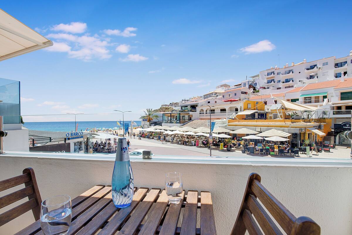 Una terraza frente al mar con bancos de madera, vista despejada de la playa y el océano, y una hilera de edificios coloridos al fondo.