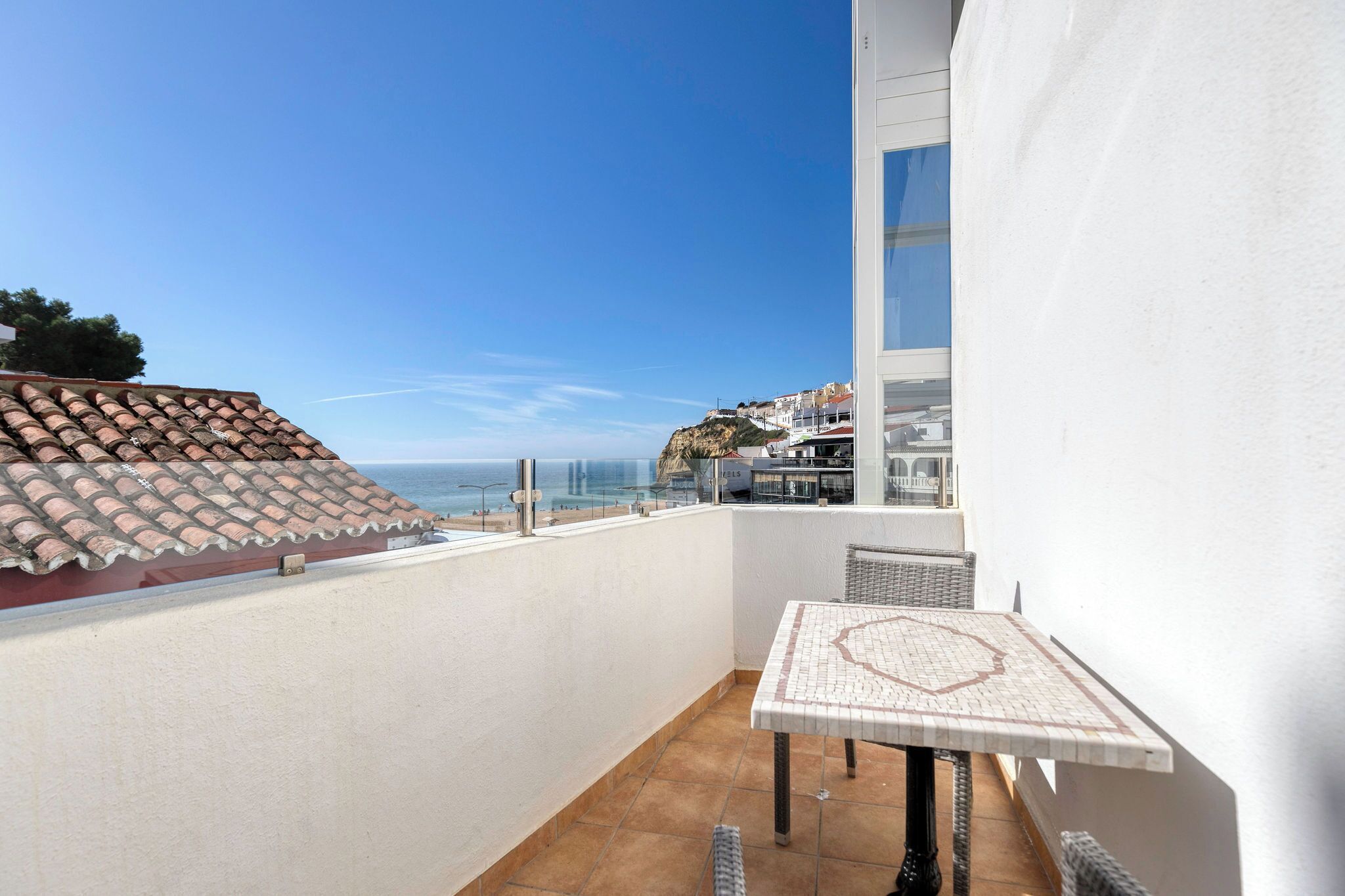 A balcony with a tiled floor, a small table with a patterned tablecloth, and a view of the sea and buildings in the distance.