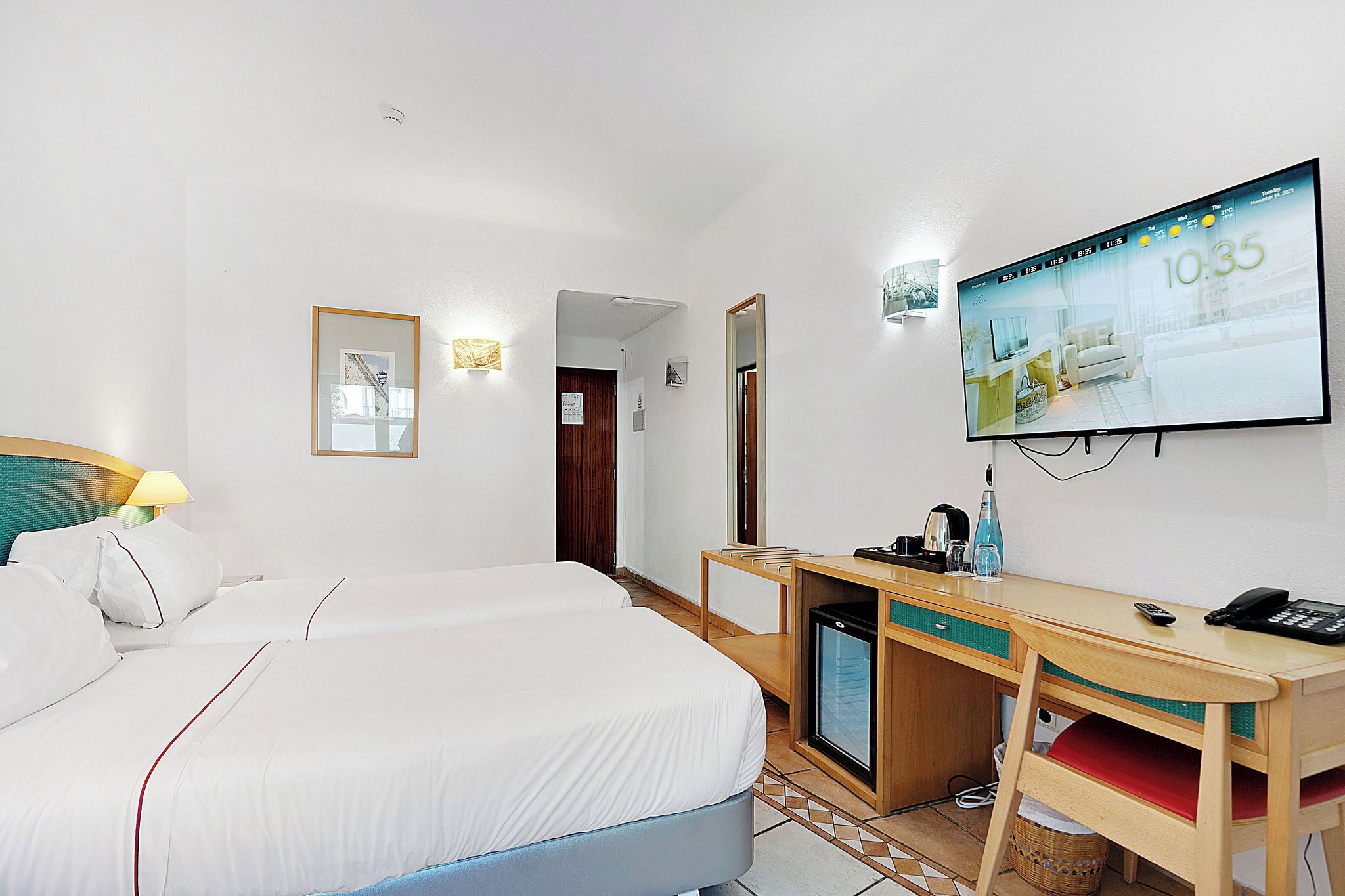A hotel room with two beds, a desk, a television, and a small refrigerator.