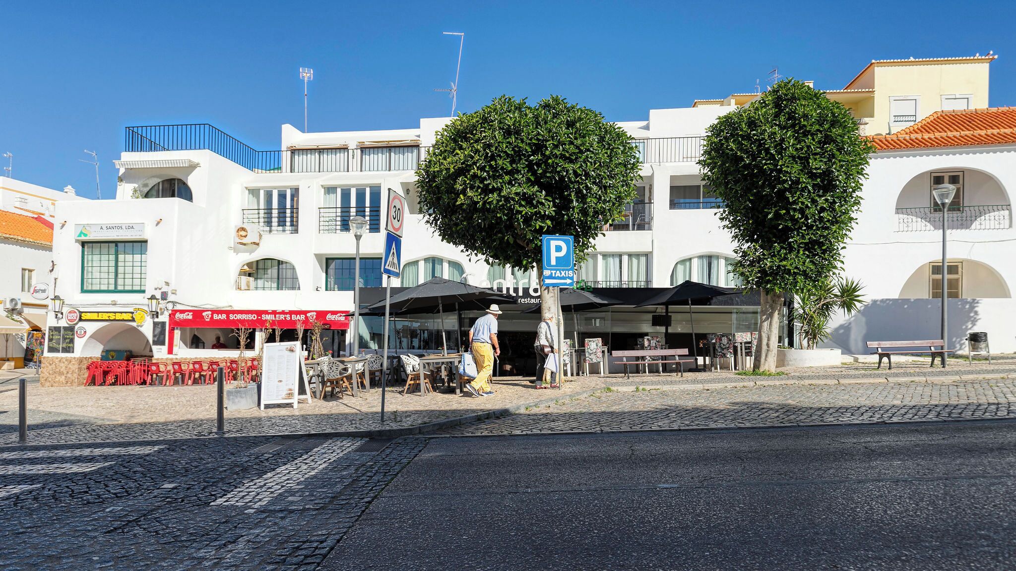 A street scene with a white building, a taxi stand, and a café named 'Sorriso - Smiler's Bar'.
