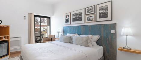 A hotel room with a bed, a desk, and a chair. There are framed pictures on the wall and a lamp on a side table.