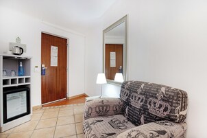 A hotel room with a wooden door, a mirror, a sofa, a lamp, a minibar, and a small kitchenette.