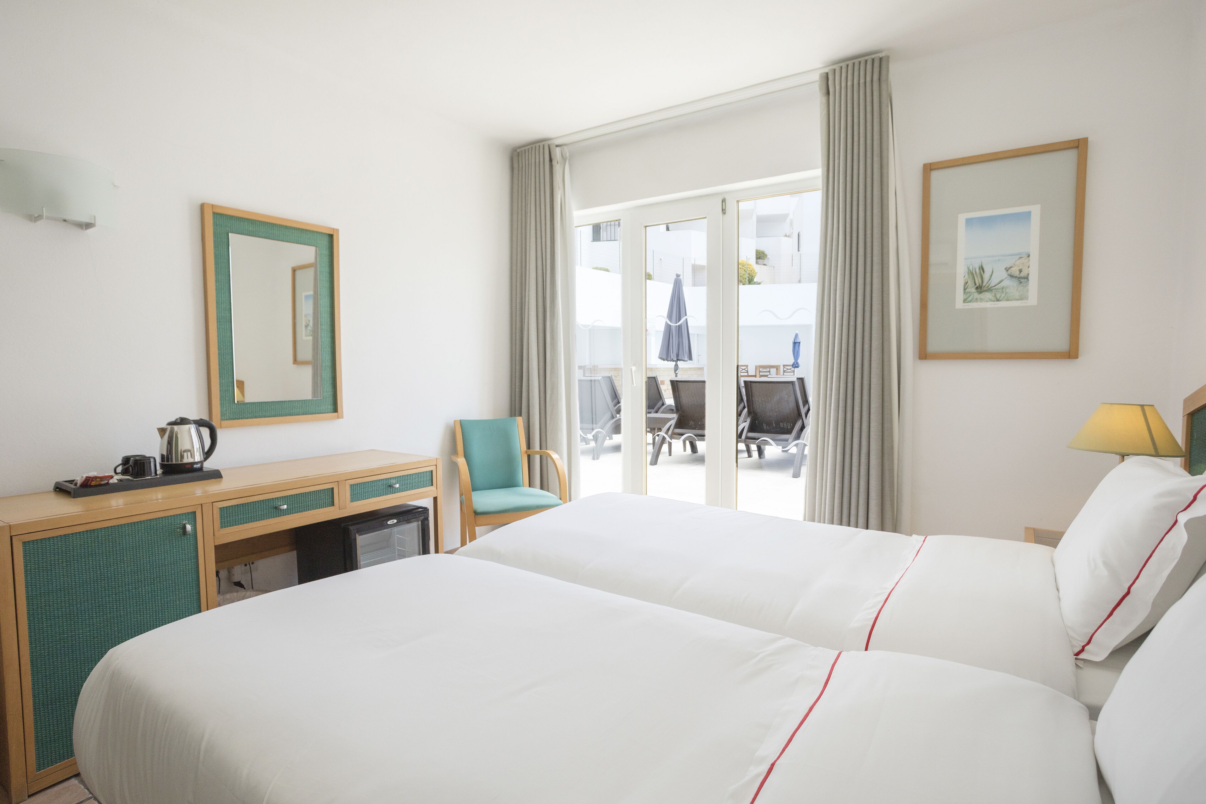 A hotel room with a bed, a desk, a chair, a small table with a kettle, and a view of the outside balcony.