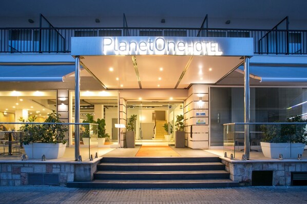 Entrada del Hotel PlanetOne con un diseño moderno, que presenta un fachada de vidrio, una escalera que conduce a la entrada y plantas en macetas alrededor del área.