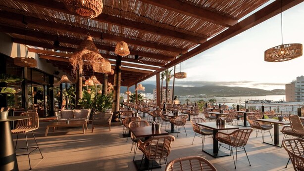 An outdoor dining area with wicker furniture, hanging wicker lamps, and a view of the water and mountains. - Globales Honolulu (Calvia)