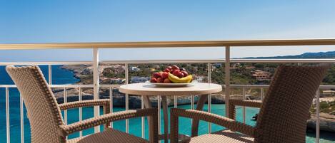 A balcony with wicker chairs, a round table with fruit, and a view of the sea and buildings.