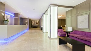 A modern hotel lobby with a reception desk, a seating area with purple sofas, and a large abstract painting on the wall. - Globales America (Manacor)