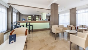A modern hotel lobby with a bar area, seating arrangements, and a view of the city through large windows. - Globales America (Manacor)