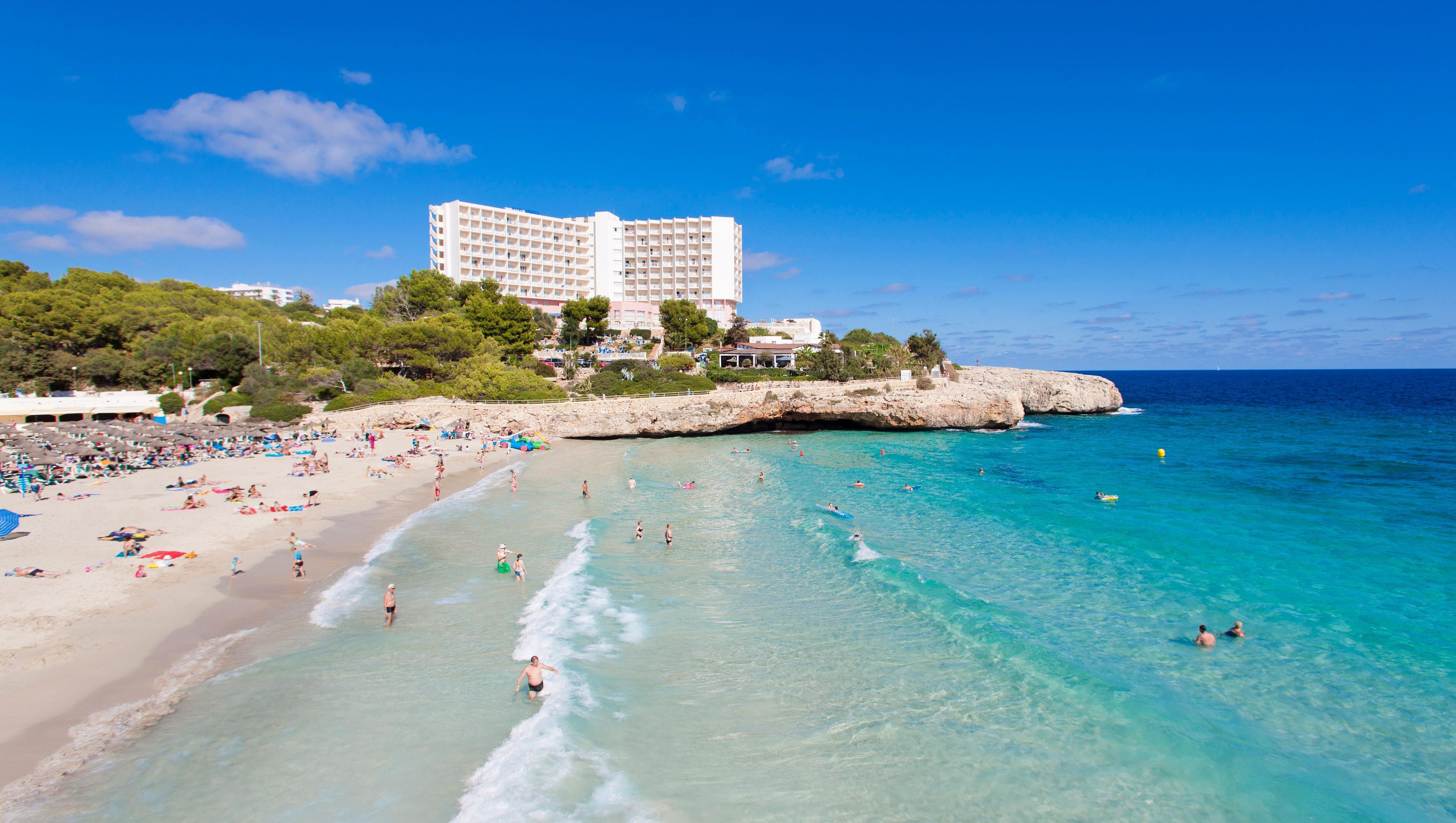 A beach with clear turquoise water, a sandy shore, and a large hotel building in the background.