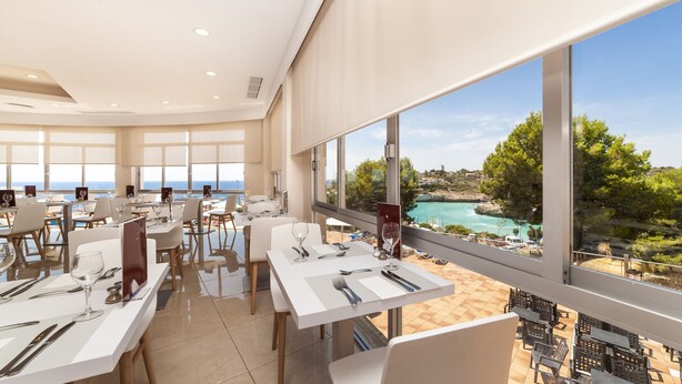 A restaurant with tables set for dining, large windows offering a view of the sea and a coastal landscape. - Globales America (Manacor)