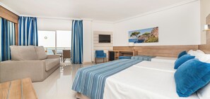 Habitació d'hotel amb un sofà, un llit amb coixins blaus, un escriptori amb una televisió i un quadre d'un paisatge.