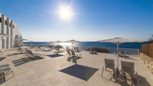A sunlit outdoor seating area with lounge chairs and umbrellas overlooking a body of water. - Globales America (Manacor)