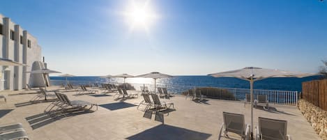 A sunlit outdoor seating area with lounge chairs and umbrellas overlooking a body of water.