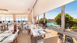 A restaurant with tables set for dining, large windows offering a view of the sea and a coastal landscape. - Globales America (Manacor)