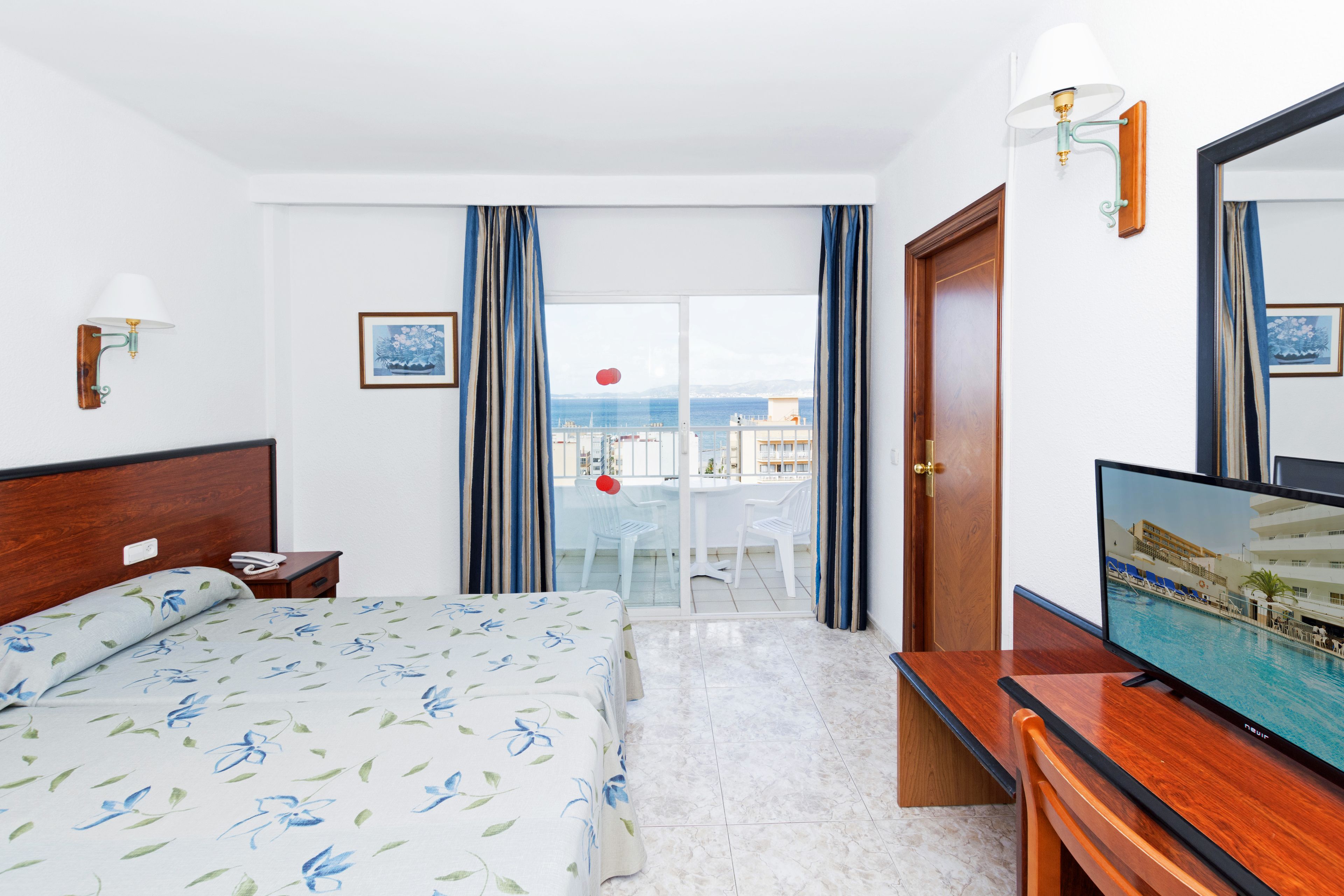 Habitación de hotel con cama, televisión, escritorio, mesita de noche, ventana con cortinas y vista al mar.