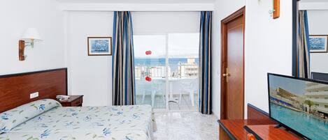 Habitación de hotel con cama, televisión, escritorio, mesita de noche, ventana con cortinas y vista al mar.
