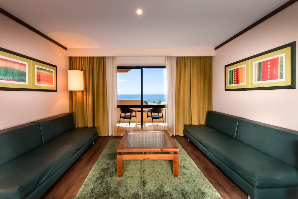 A hotel room with two green sofas, a glass-top coffee table, a view of the sea, and framed artwork on the walls.
