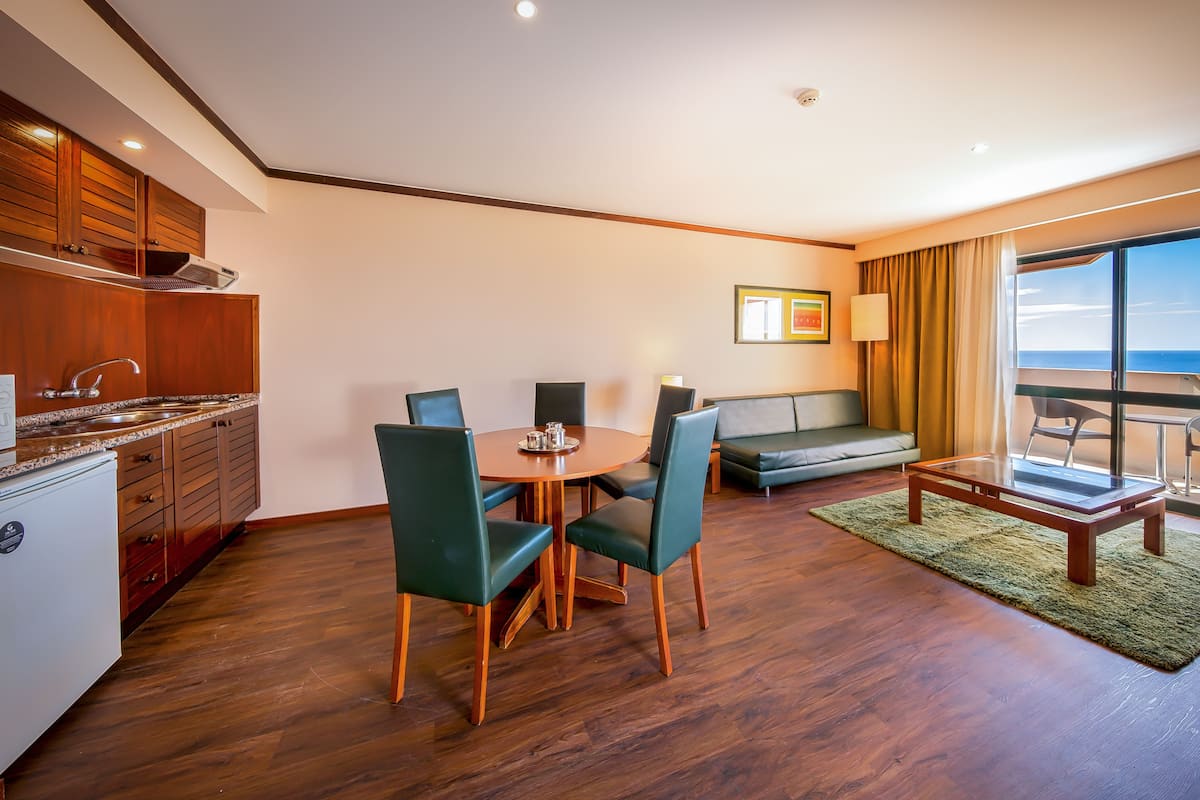 A modern hotel room with a dining area, a sofa, a balcony with a view of the sea, and a kitchenette.