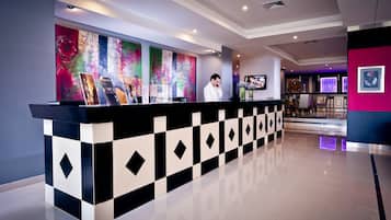A modern hotel reception area with a check-in counter, a large abstract painting, and a bar area in the background.